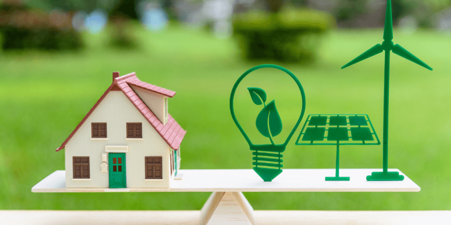 Scale on a table with house on one side and energy sources on the other, lightbulb, solar panel and windmill